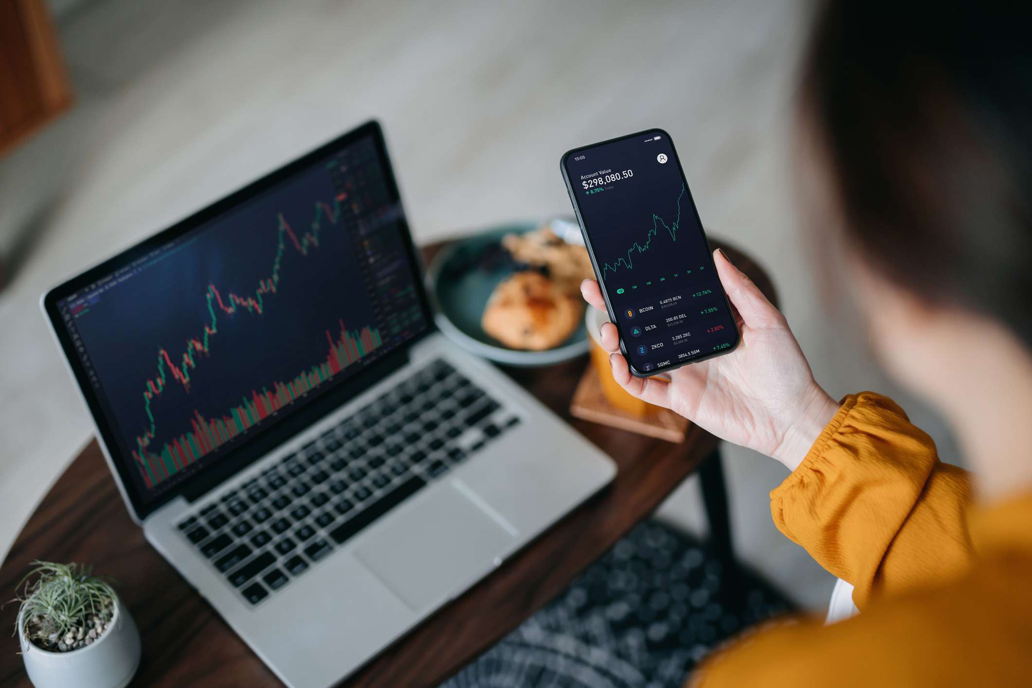 Over the shoulder view of Asian woman holding smartphone, analyzing investment trading data on crypto graph with smartphone and laptop while working at home. Cryptocurrency, bitcoin, digital ledger