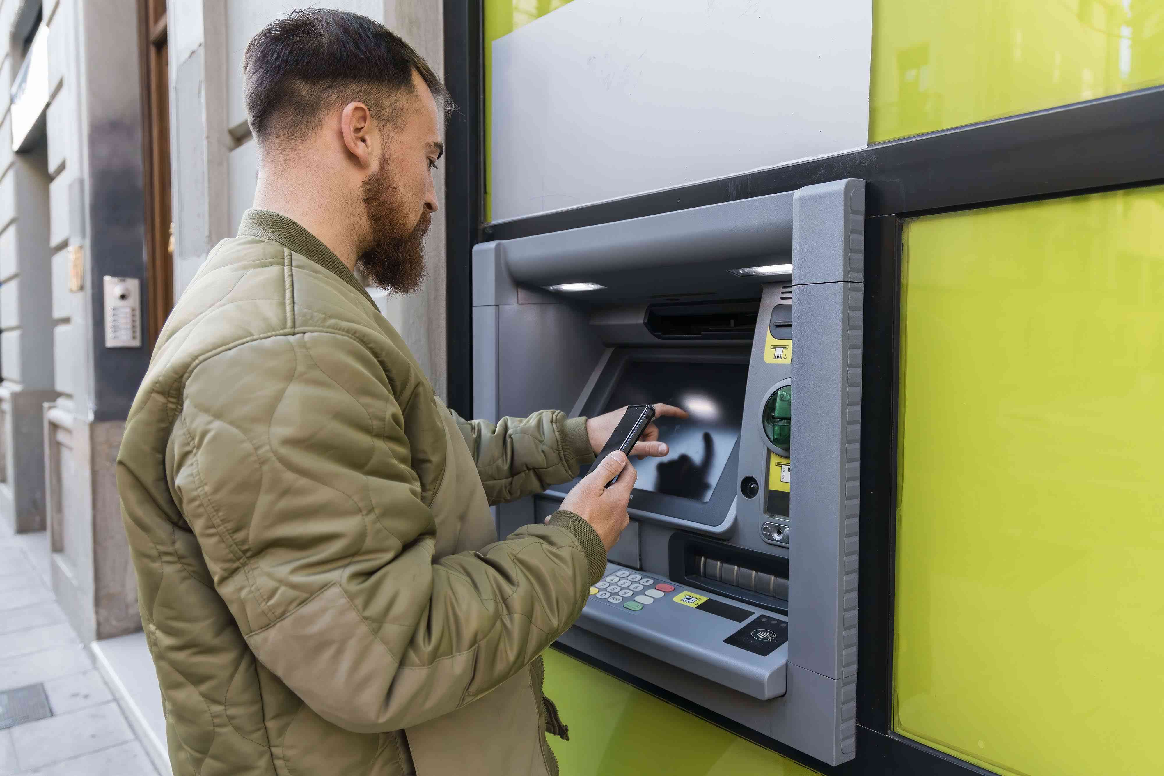 Young man with smart phone using ATM